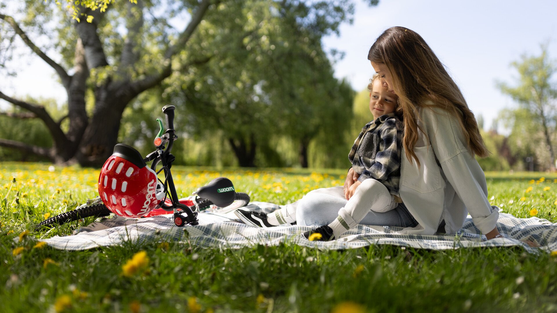 Een jongetje en zijn moeder zitten op een picknickkleed in het park naast een woom-kinderfiets.