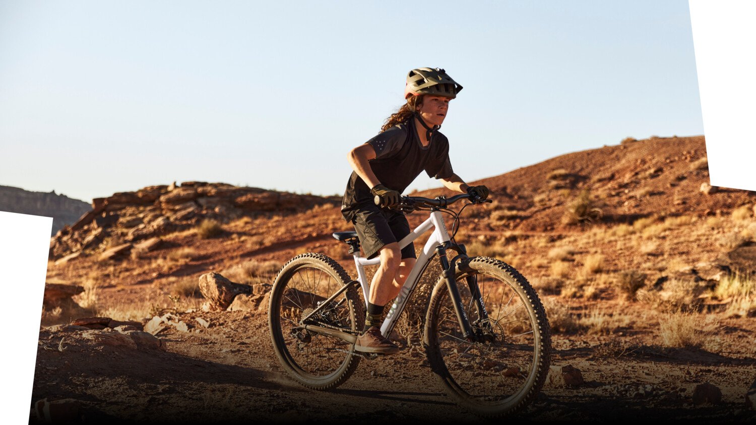 Junge mit Fahrradhelm auf dem Kopf fährt auf einem weißen Mountainbike mit Federgabel von woom auf einem ruppigen Trail.