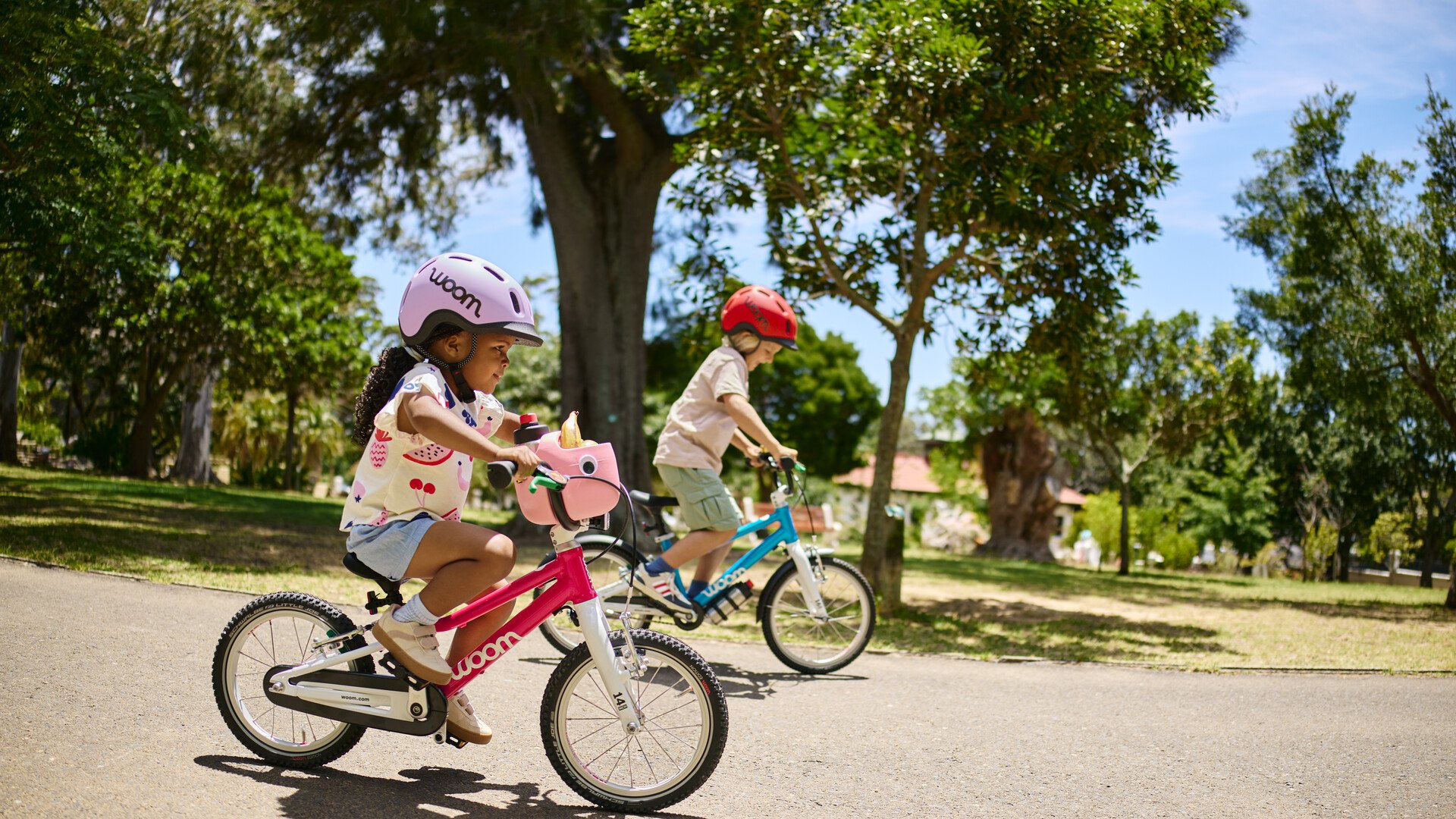 Zwei Kinder mit dem READY Helm auf dem Kopf fahren auf Fahrrädern von woom durch einen Park.
