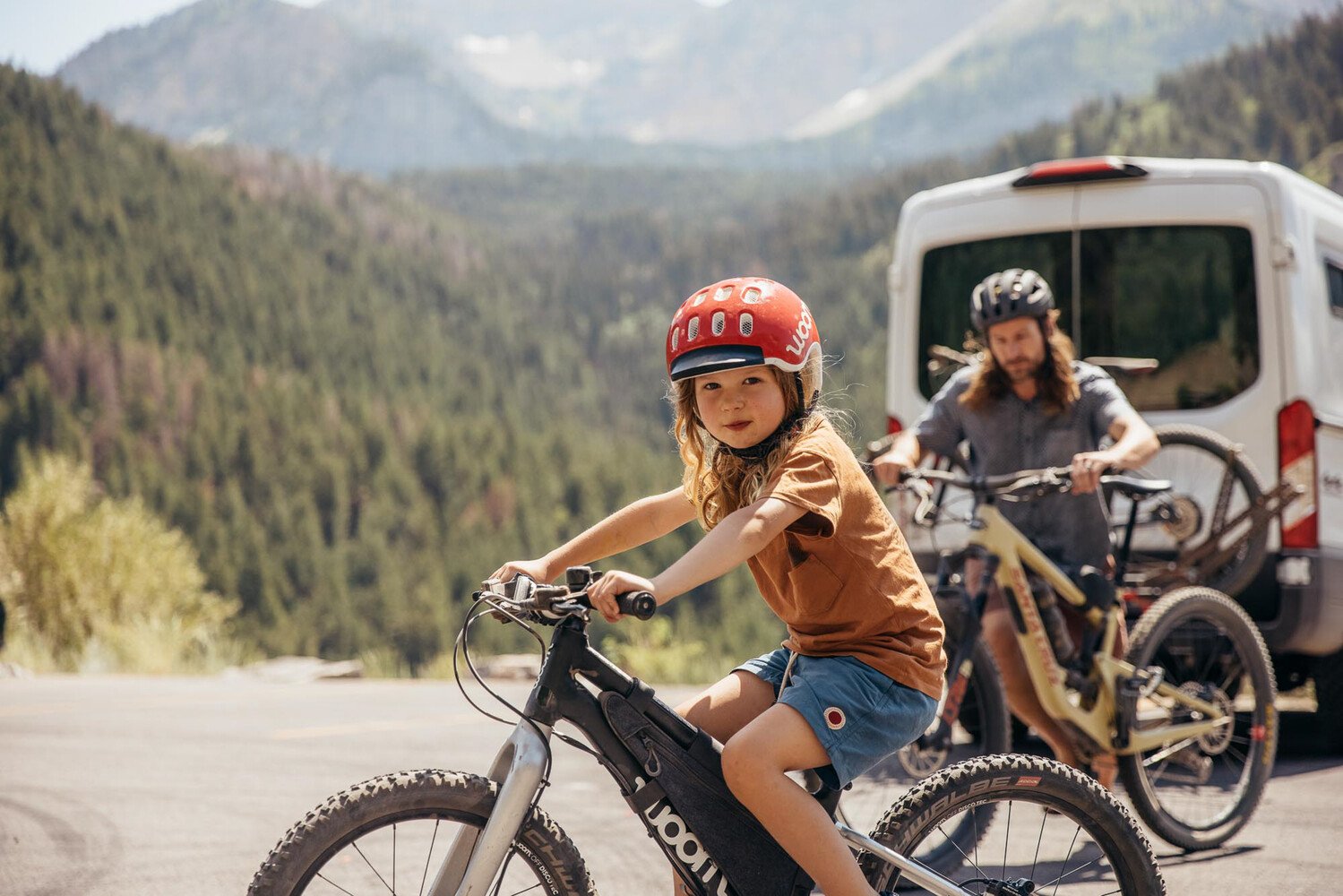 Auf einem schwarzen woom OFF Mountainbike sitzt ein Bub mit rotem Radhelm auf dem Kopf, im Hintergrund sind Bäume und Berge.
