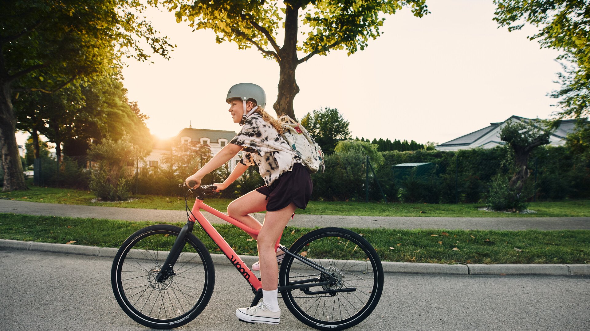 Mädchen mit Fahrradhelm auf dem Kopf fährt mit einem orangen woom EXPLORE Fahrrad im Sonnenuntergang auf der Straße.