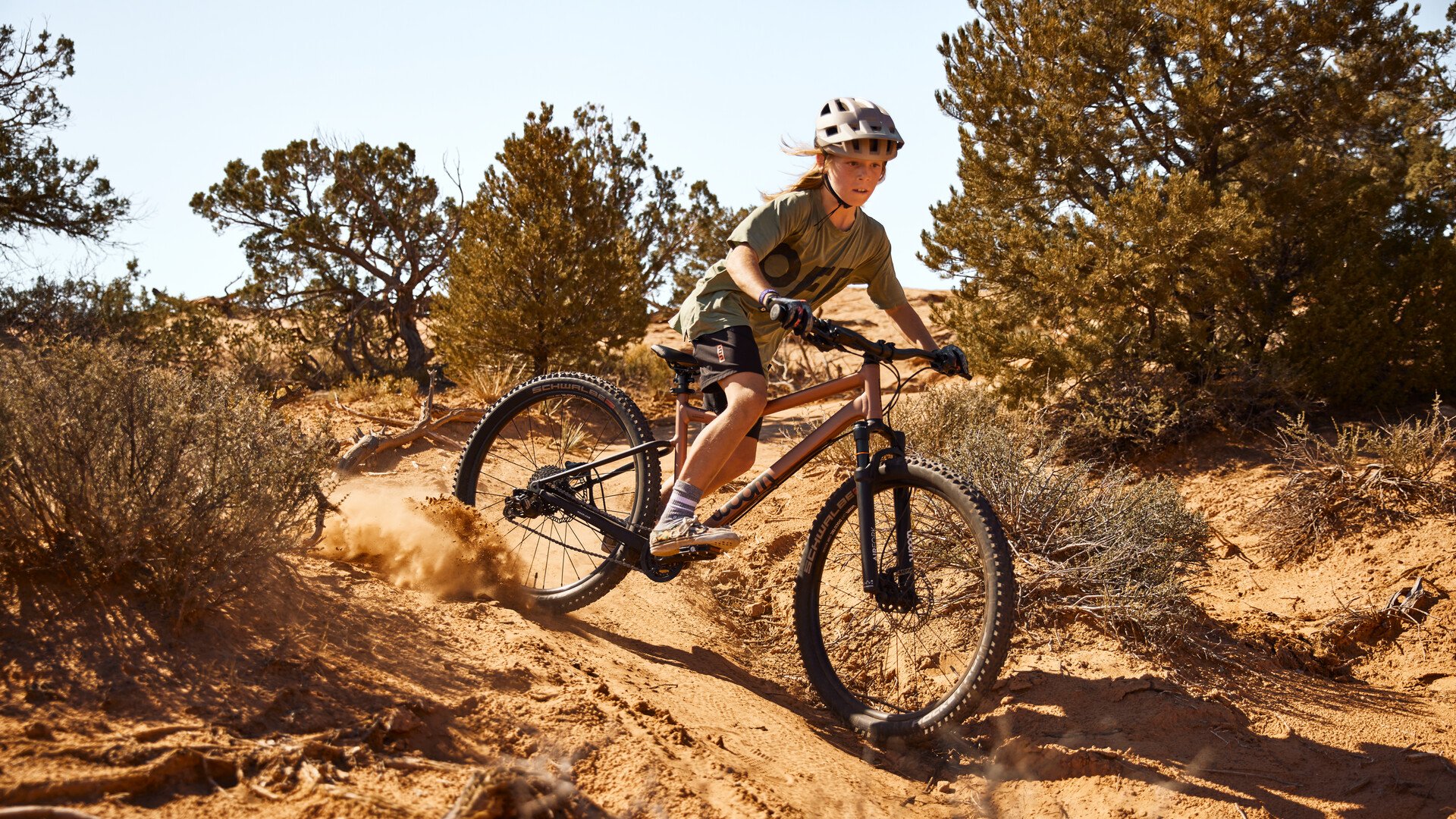 Junge mit Fahrradhelm auf dem Kopf driftet mit einem kupferfarbenen woom OFF AIR Mountainbike auf einem sandigen Trail.