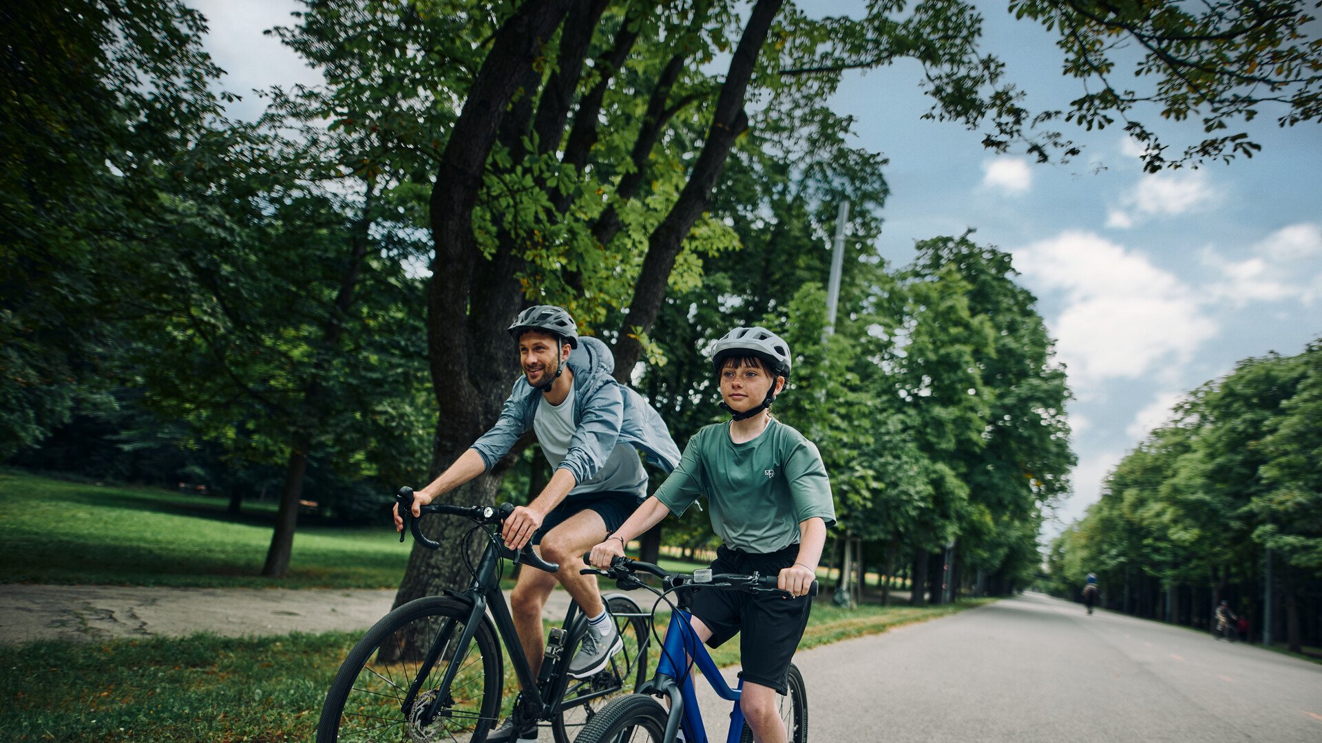 Junge mit Fahrradhelm auf dem Kopf fährt begleitet von seinem Vater auf einem blauen woom EXPLORE Fahrrad im Grünen.