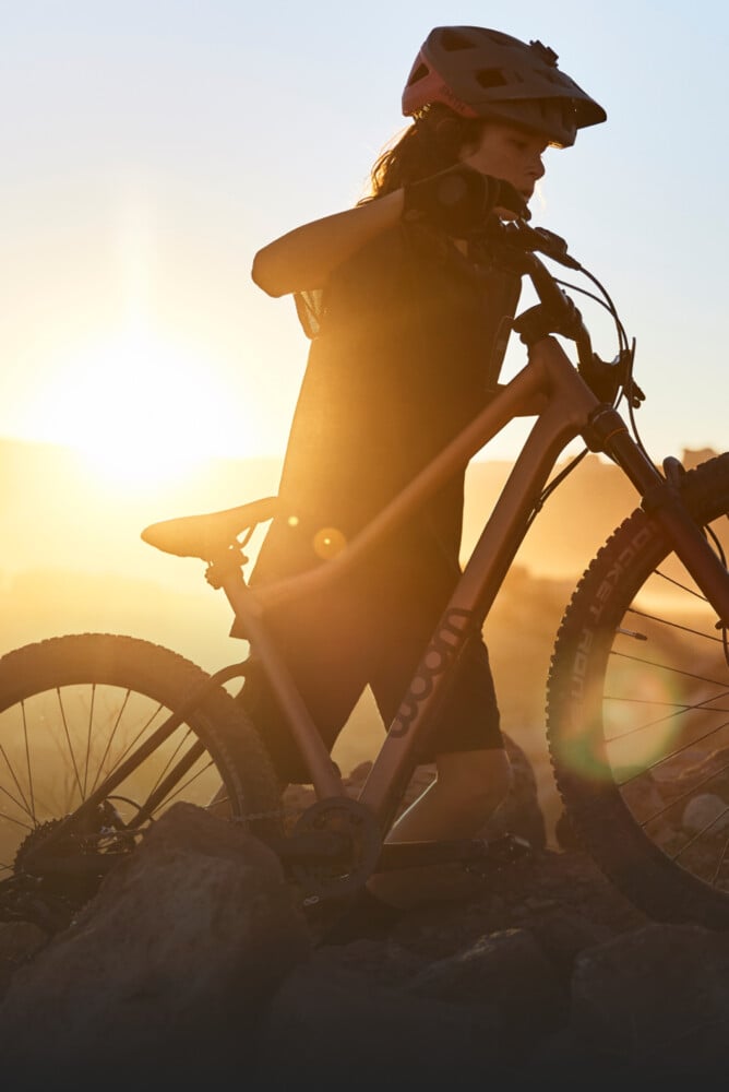 En tonåring med cykelhjälm skjuter sin mountaincykel woom OFF uppför en sluttning i solnedgången.