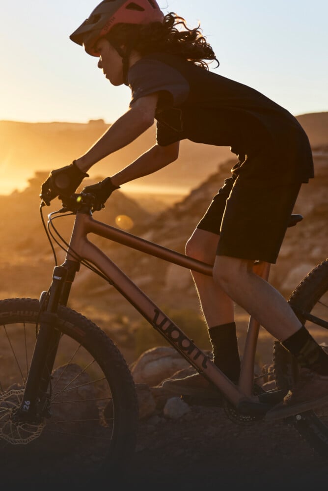 Kind mit Fahrradhelm auf dem Kopf fährt bergab in der Grundposition auf einem woom OFF AIR Mountainbike im Sonnenuntergang.