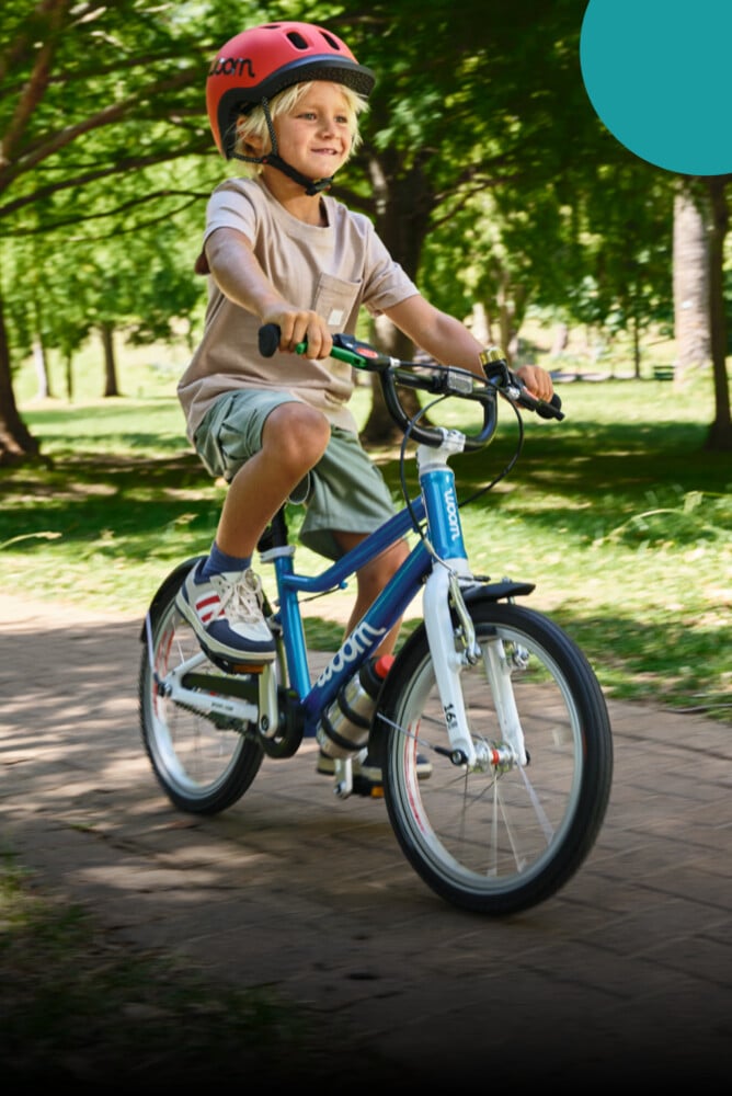 På sadeln till en blå woom GO 3-cykel sitter en pojke med röd READY-barnhjälm på huvudet.